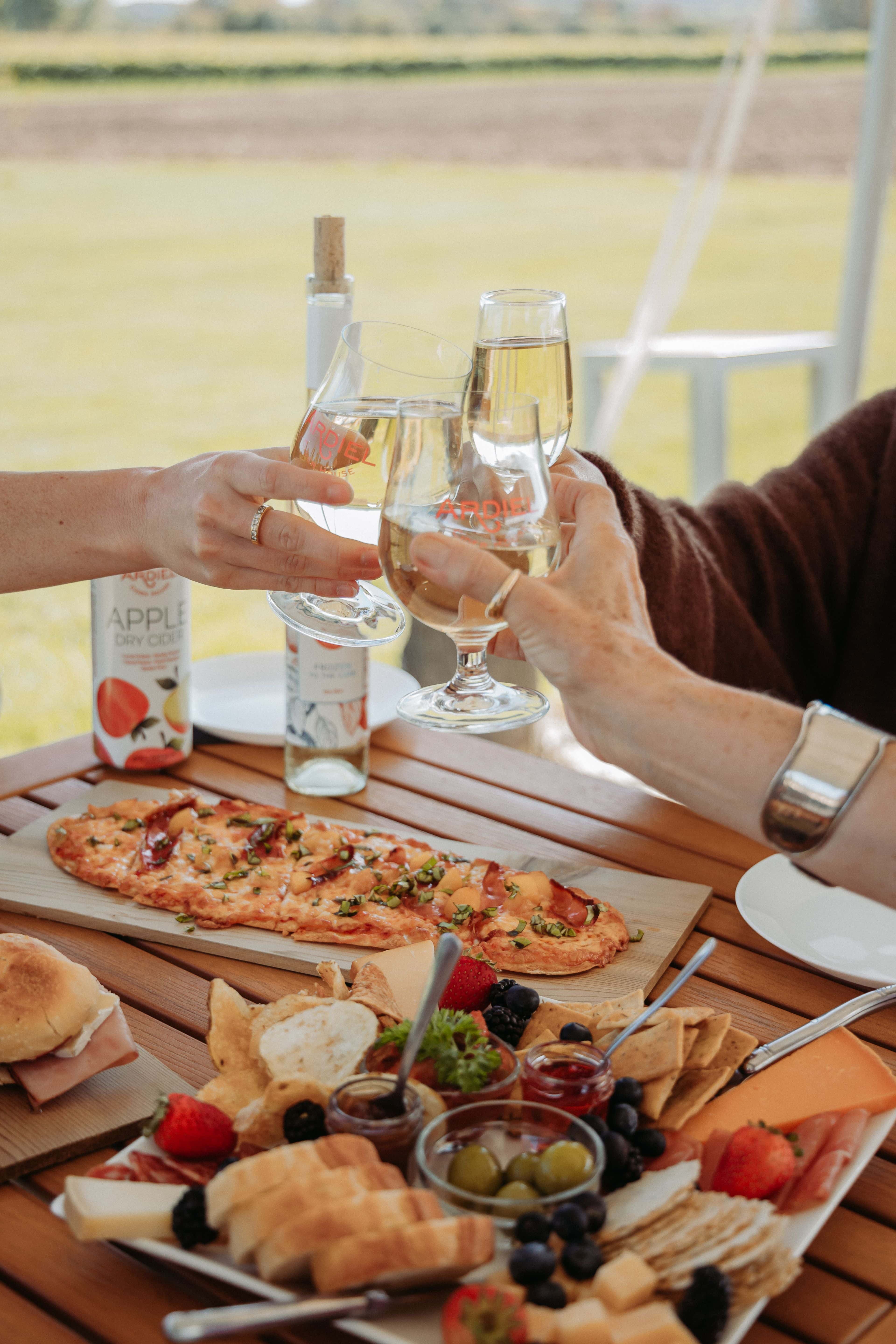 Three friends clink their glasses together above a table full of colourful dishes, sitting on the outdoor patio at Georgian Hills Vineyards in Clarksburg