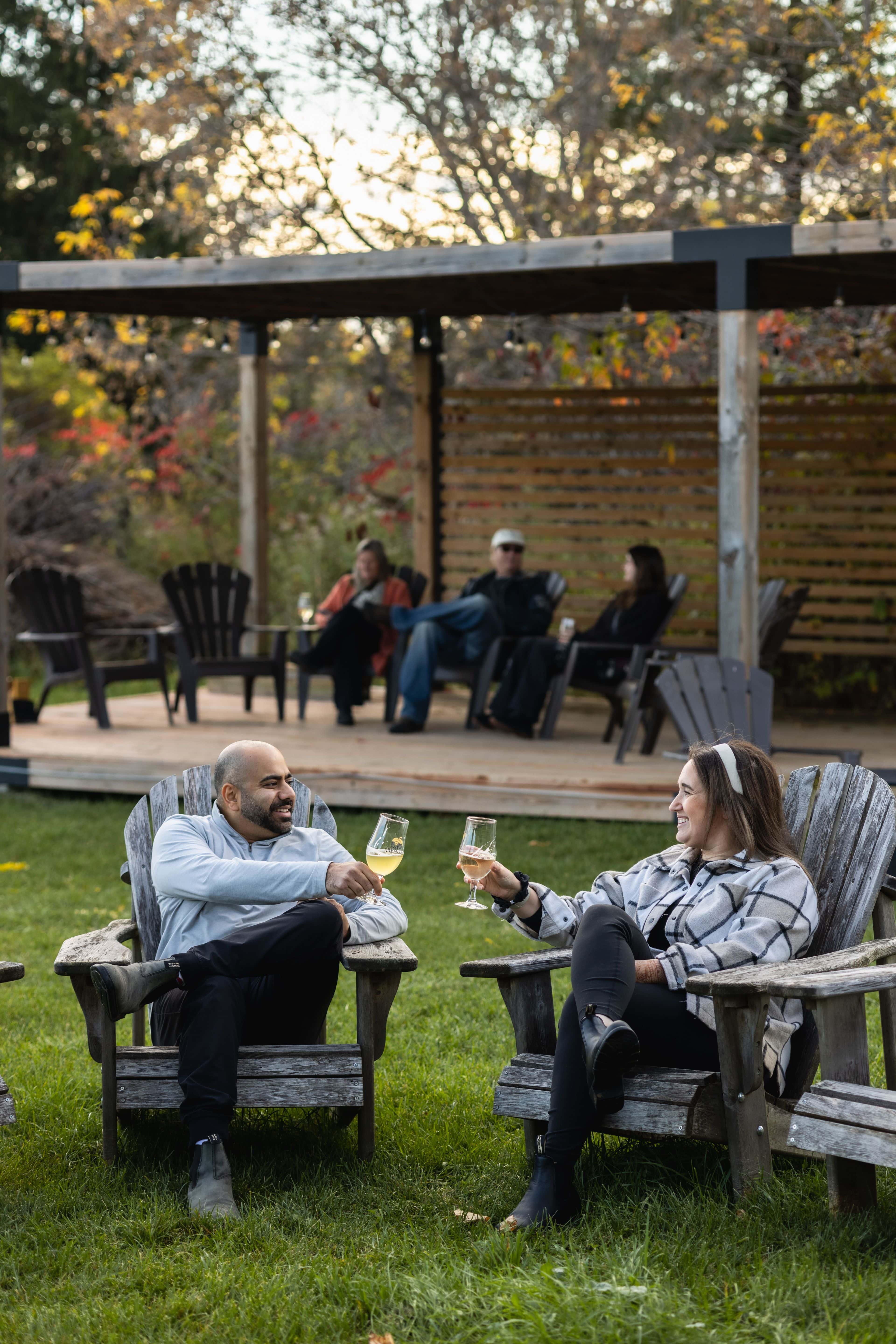 A couple sitting in muskoka chairs cheersing their glasses together as they enjoy local sips at Grey & Gold Cider on a cool evening in Fall