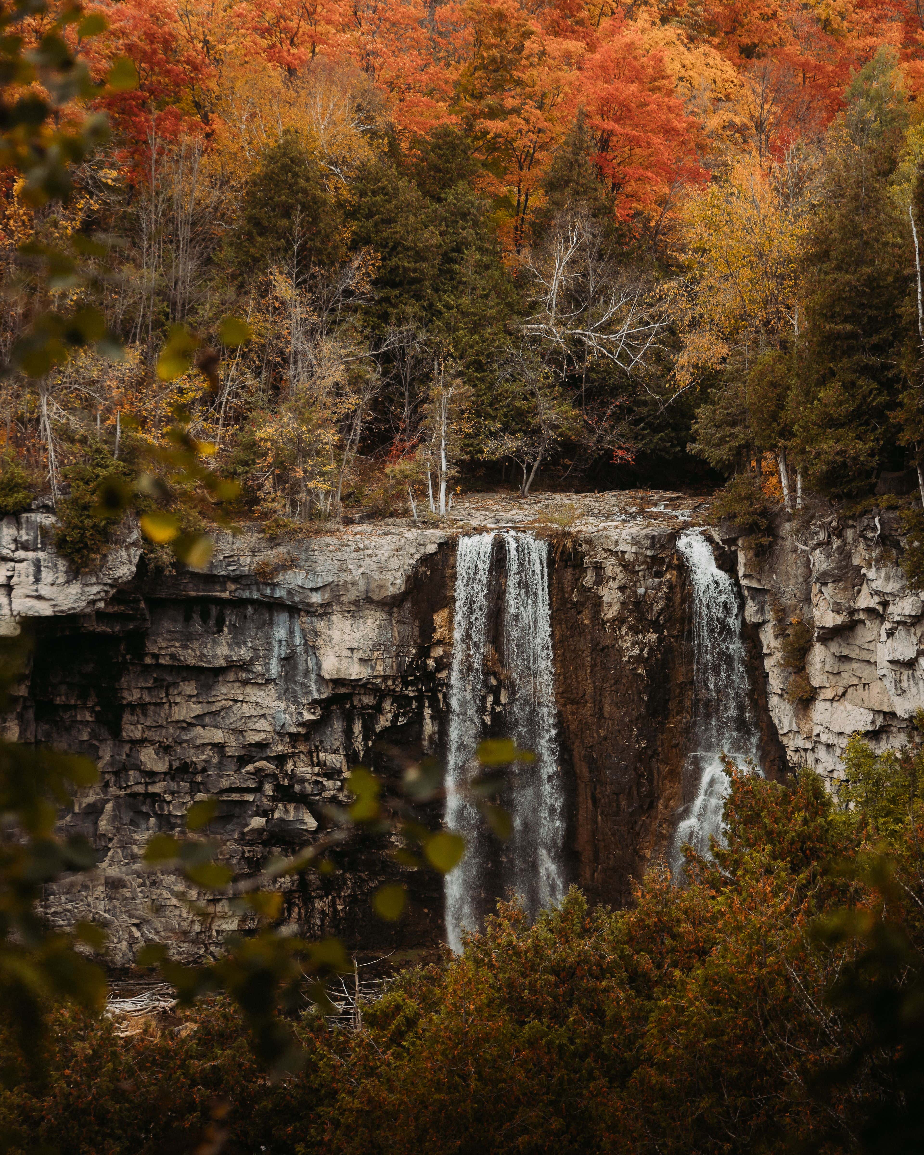 Natural waterfall flowing over the rock cliffs, surrounded by a forest of amber and red leaves on a Fall afternoon.