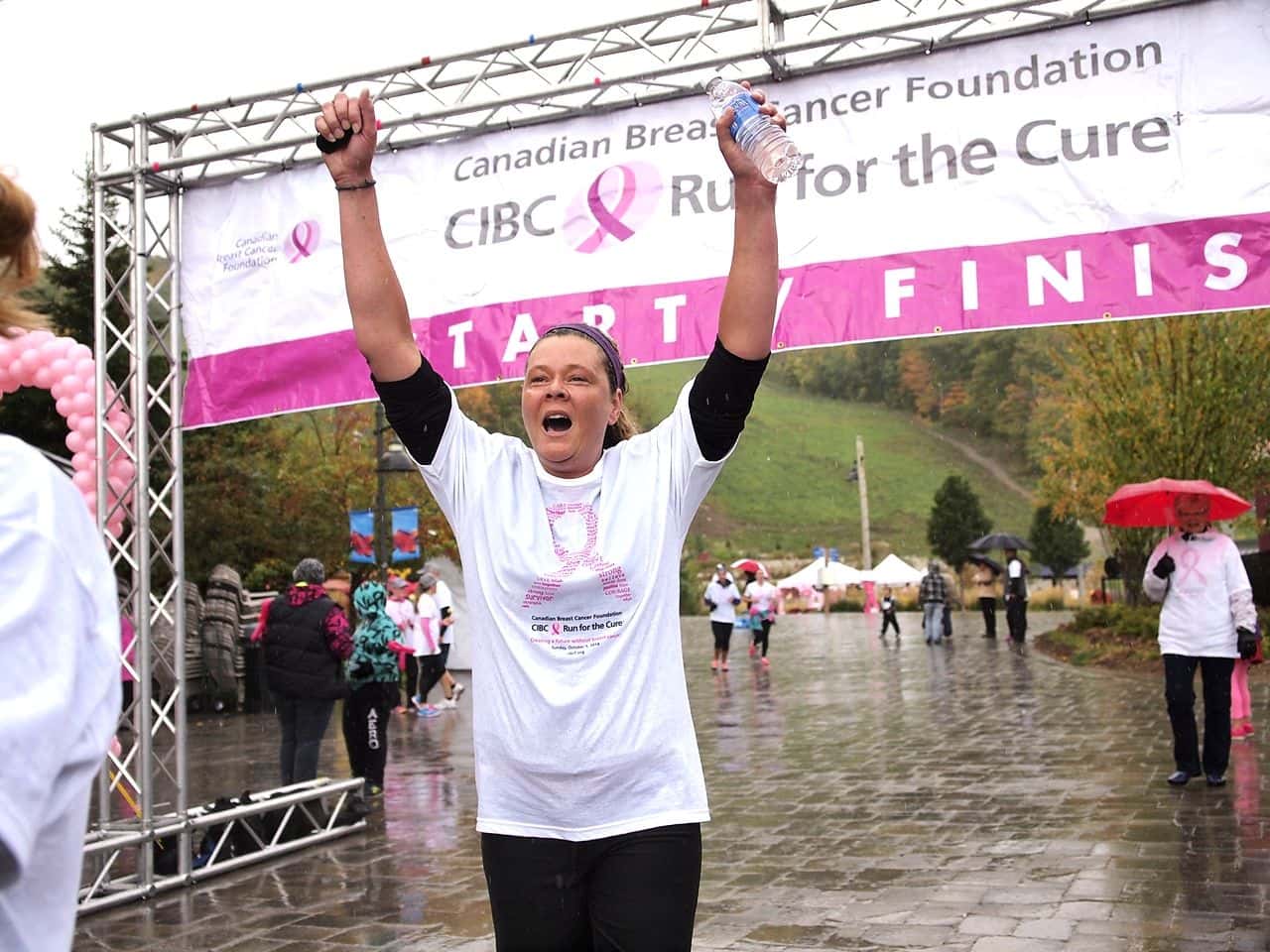 Woman running with her hands in the air, yelling with excitement as she crosses the finish line for CIBC Run for the Cure on a rainy day in the Blue Mountain Village Events Plaza.