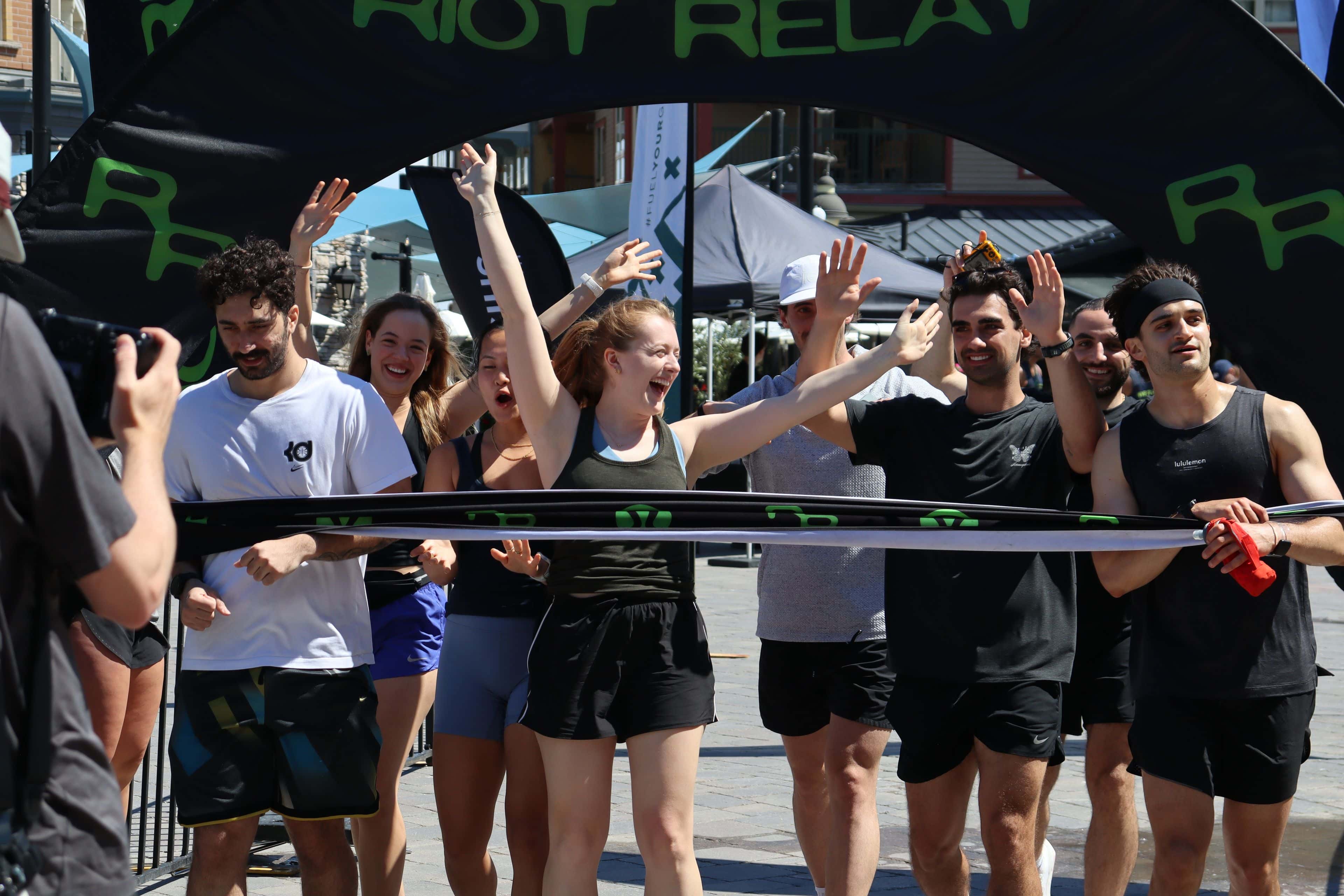 A group of racers cheer with hands in the air as they make it across the finish line after running the Riot Relay Race to Blue Mountain Village.