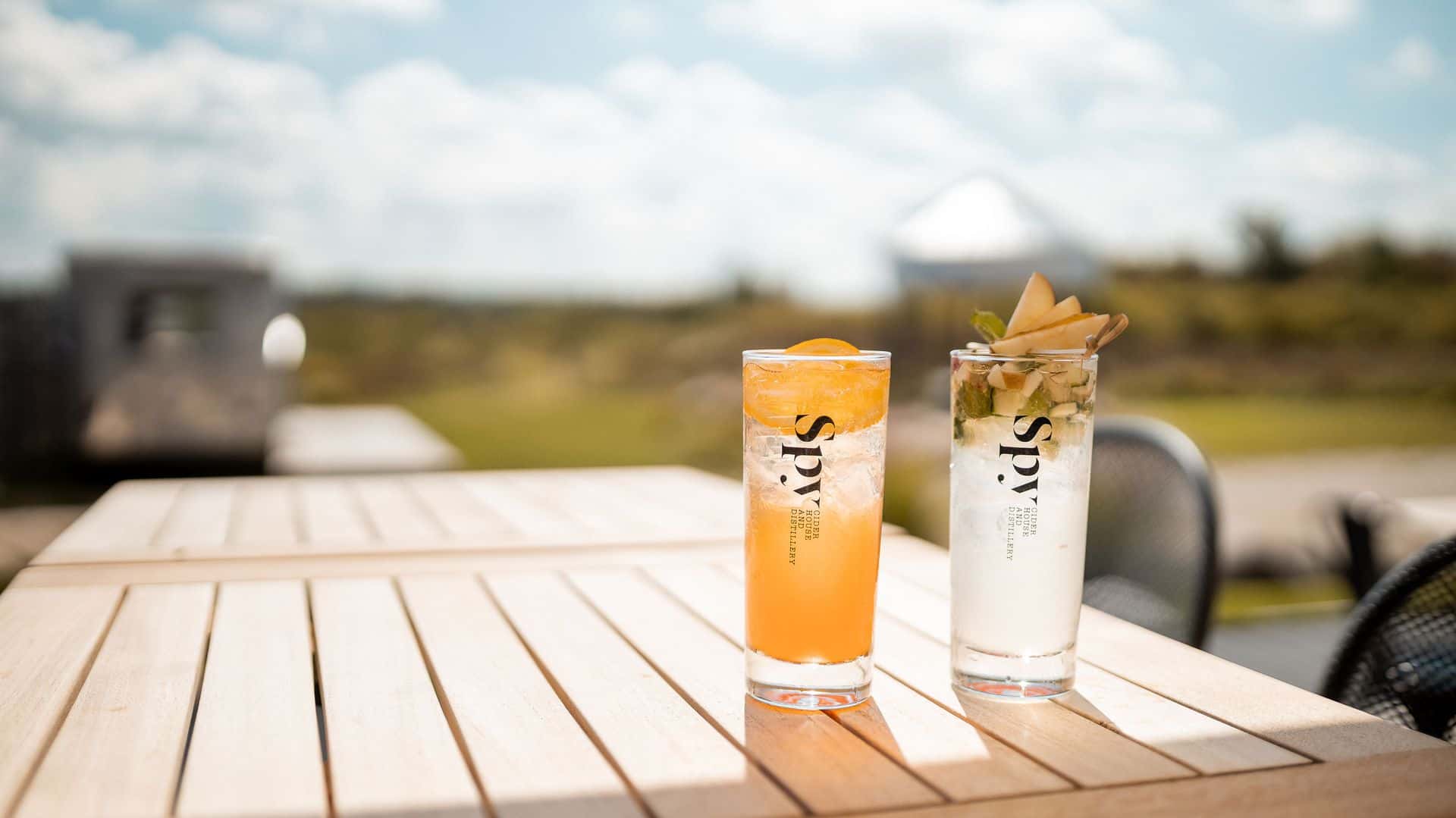 An image of two drinks from Spy Cider + Distillery on a table outside during summer.