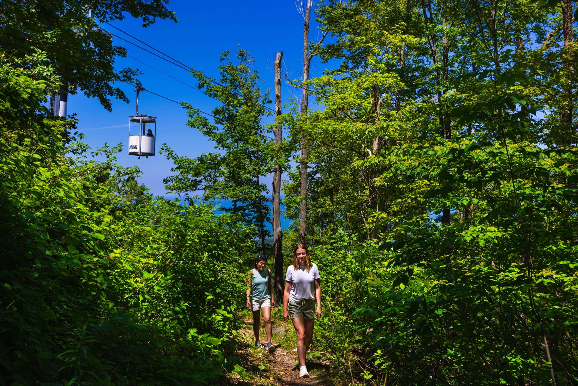 Two girlfriends hiking up the Blue Mountain Resort Trails on a blue bird day with the open-air gondola ascending in the background